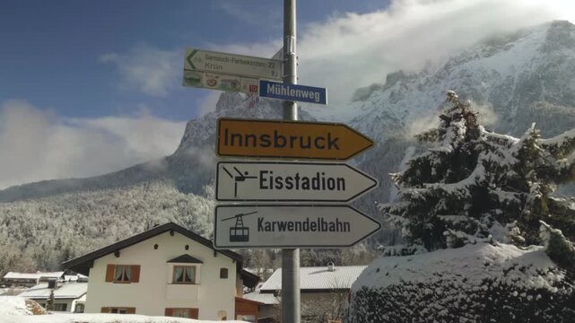 Road signs in Mittenwald, Bavaria, Germany, pointing toward Innsbruck and local destinations. Snow covered houses and navigation signs stand against Karwendelgebirge, with Westliche Karwendelspitze