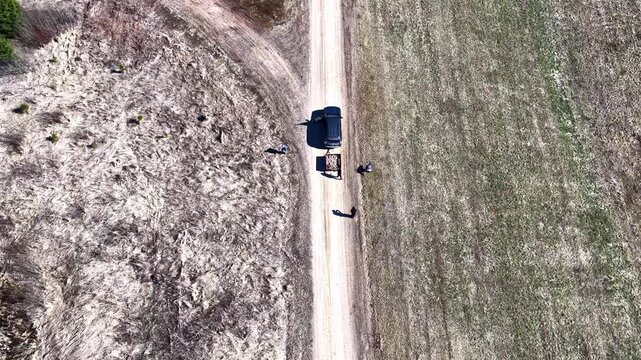 Top-down drone view of people beside a parked vehicle on a rural dirt road, dividing pale field and brushy terrain in an abstract countryside composition.