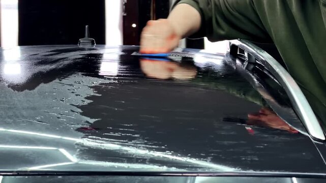 Close-up of a hand applying detailing product across a glossy black car roof with a blue applicator pad, showing paint care and professional auto detailing.