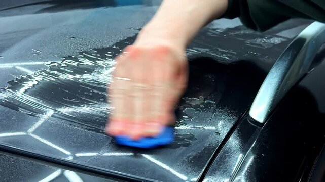 Close-up of a hand applying product to a black car hood with a blue applicator pad, showing vehicle detailing and paint care in action.