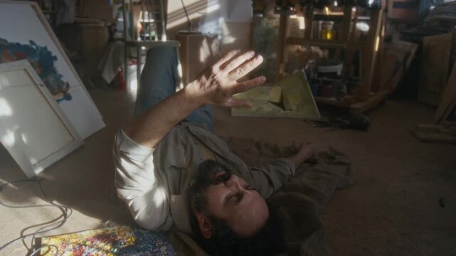 Depressed male artist lying on messy floor surrounded by canvases, looking at his palm in bright sunlight while recovering from emotional breakdown