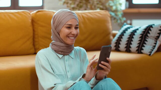 A young woman with light brown skin and a hijab sits on the floor of her living room. She is smiling and texting on her smartphone while sitting against a bright yellow couch.