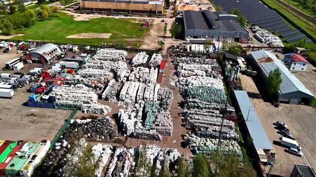 Aerial flyover above vast plastic recycling factory yard in Jelgava, Latvia showing massive stockpiles of sorted plastic bales and industrial buildings