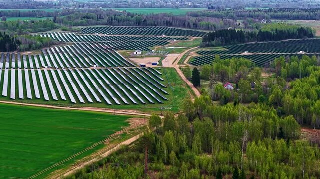 Aerial panning shot over large solar panel farm with substation, access roads, spring birch forest patches, and surrounding green fields in rural Latvia