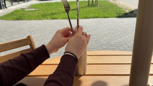 Woman taking and placing knife and fork into cutlery holder with napkins on outdoor restaurant table service, and casual cafe atmosphere in sunlight. 