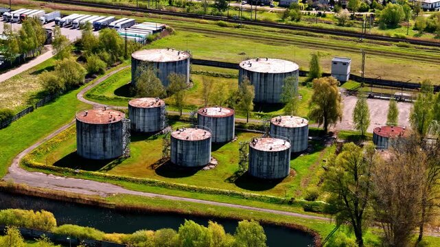 Aerial parallax view of large cylindrical industrial storage tanks surrounded by green trees, canal waterway, and railway in Latvia