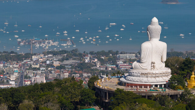 Aerial view of Big Buddha Phuket statue overlooking the bay with numerous boats, urban buildings, and lush green trees Karon, Phuket, Thailand.