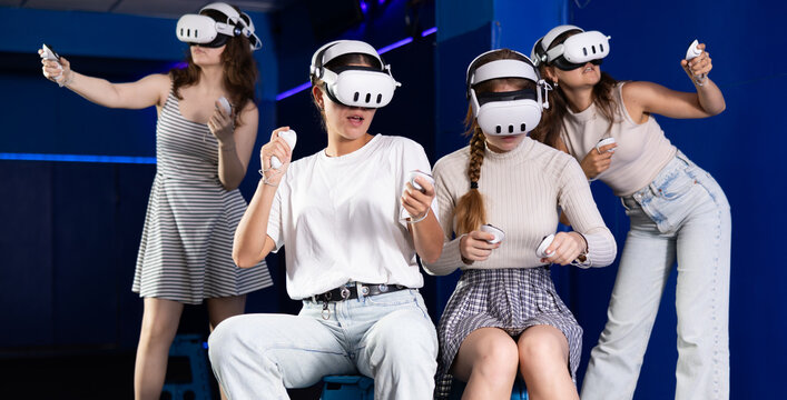 Group of cheerful excited young female friends having fun with virtual reality game during bachelorette party, wearing VR headsets and holding controllers in blue-lit room
