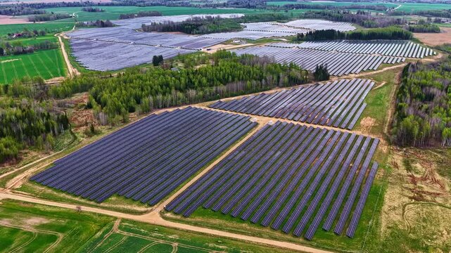 Aerial view of large solar panel farm with multiple sections separated by forest patches and surrounded by green agricultural fields in Latvia