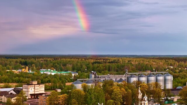 Aerial view of dramatic rainbow over grain elevator silos and historic church surrounded by autumn forest in Jelgava, Latvia under moody skies