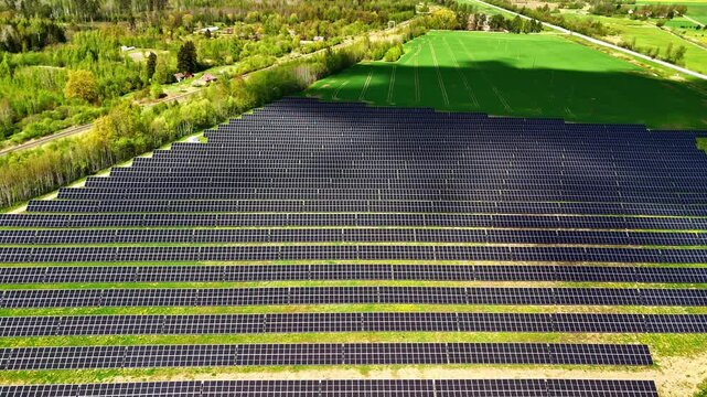Aerial flyover above solar panel farm in rural Latvia with green agricultural field, spring trees, and farm buildings visible in surrounding countryside