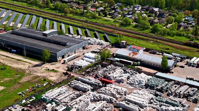 Aerial view of plastic recycling factory with solar panel arrays and massive plastic bale stockpiles as freight train passes on nearby railway in Latvia