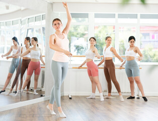 Obraz premium Expressive solo dance moment in ballet rehearsal. Confident young female dancer performing graceful movements in sunlit studio in front of group practicing at barre