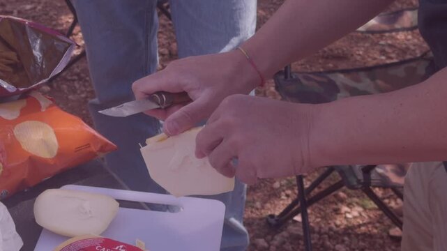Hands slicing wedge of cheese with knife during outdoor camping picnic preparation in nature