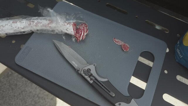 Cured sausage slices and folding knife resting on cutting board near grill