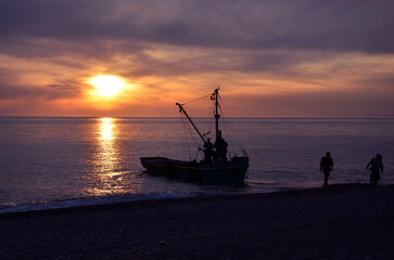 Obraz premium Fishermen returning to shore with their boat at sunset. Silhouette of sailors and fishing vessel on the beach at dusk