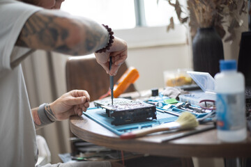 Fototapeta premium Person repairs a small electronic device on a table at home in daylight while tools and materials are nearby