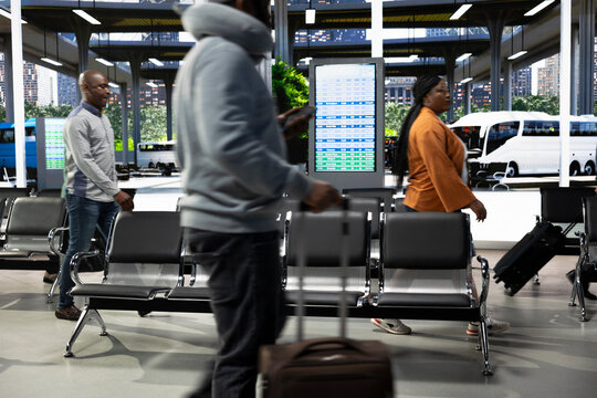Crowded bus terminal with travelers preparing for interstate travel. Passengers checking departure board schedule on screen with luggage and trolley bags before boarding long-haul journey.