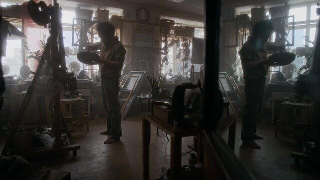 Wide shot of depressed painter putting away palette and brush, sitting on floor of his messy studio, drinking alcohol and staring at canvas while struggling to find inspiration
