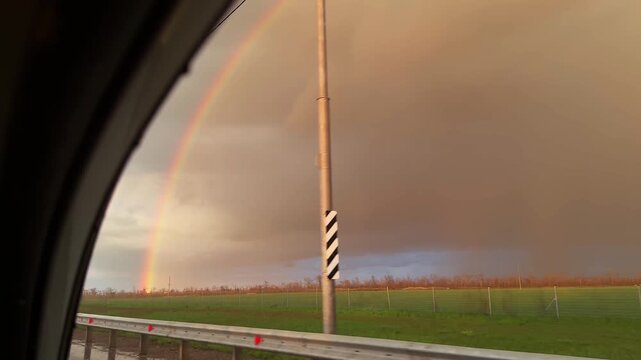 View from moving car through side window showing double rainbow over countryside landscape. Scenic roadside with guardrail and green fields under dramatic sky. Concept of travel, weather and journey