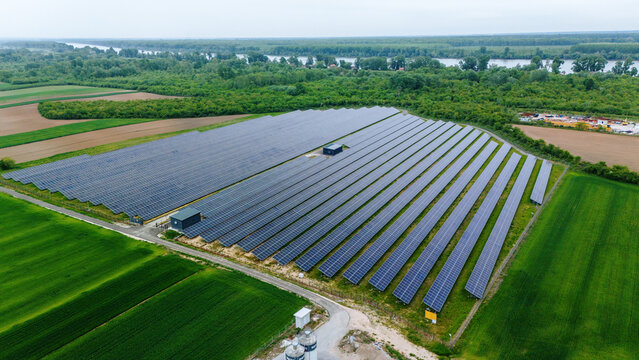 Aerial view of a large solar panel array surrounded by lush green agricultural fields and dense forest near a river in Sabac, Vojvodina, Serbia.