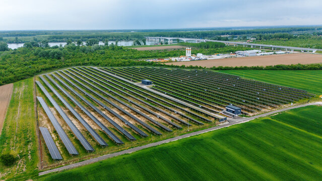 Aerial view of a large solar farm with rows of photovoltaic panels surrounded by green fields and a river with a bridge in the distance Sabac, Vojvodina, Serbia.