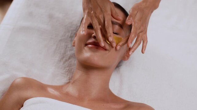A woman with light skin enjoys a facial treatment at a spa. A service provider applies gold patches under her eyes. The setting is bright and inviting.