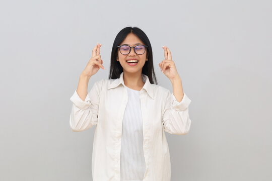 A smiling young Asian woman wearing casual clothes, crossing her fingers with her eyes closed, raising her hand, posing isolated on a white background.