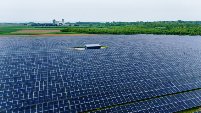 Aerial view of a vast solar panel farm surrounded by green agricultural fields and industrial buildings under an overcast sky in Sabac, Vojvodina, Serbia.