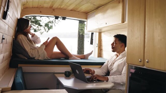 Young mixed race couple enjoying van life. The man is a freelancer working on a laptop while his girlfriend relaxes on the bed using a smartphone