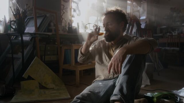Frustrated male artist sitting on floor in messy studio filled with sunlight, drinking alcohol from bottle, then throwing away destroyed artwork and suffering from deep professional crisis
