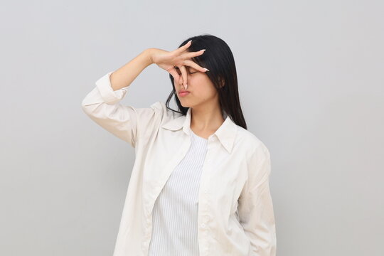 A young Asian woman in casual clothes, covering her nose with her hand because of a bad smell, stands alone on a white background.