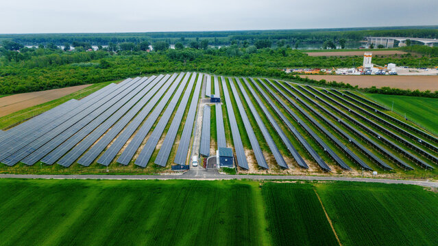 Aerial view of a large solar panel farm surrounded by lush green agricultural fields and dense forests near a river in Sabac, Vojvodina, Serbia.