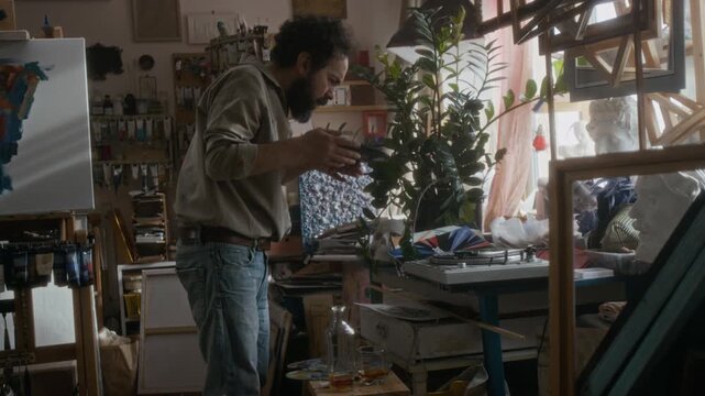 Artist choosing vinyl record, blowing dust off it, playing music on vintage turntable and having whiskey during break in cozy creative studio filled with paintings and craft supplies