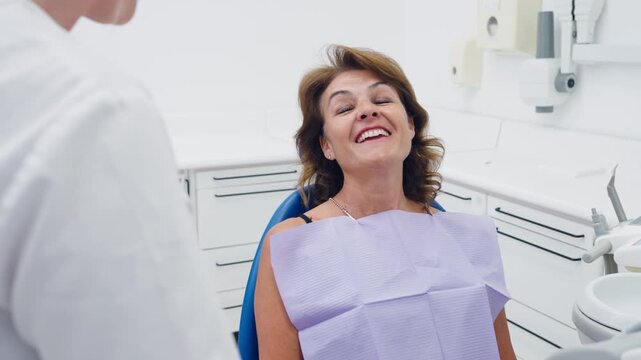 Mature female patient with a dental bib sitting in a chair, smiling and talking with her dentist in a bright, modern dental clinic