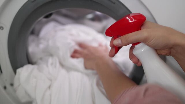 Hand holding spray bottle applies cleaning solution to white fabric inside washing machine, preparing to remove stains from laundry in a bright indoor setting