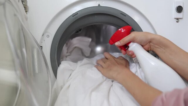 Woman applying stain remover spray to white fabric inside washing machine, demonstrating effective cleaning technique for laundry care in a bright indoor setting