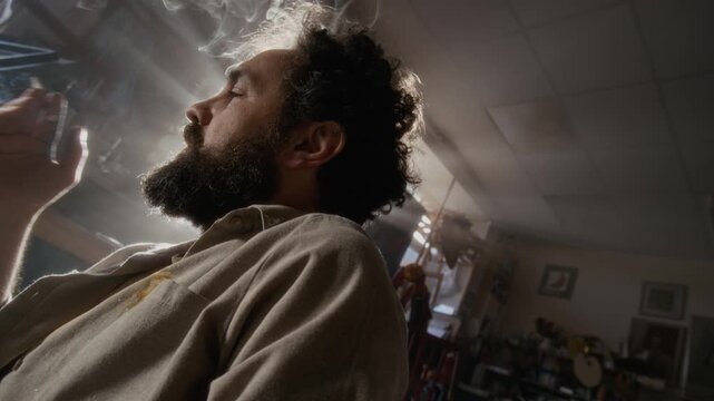 Low angle view of depressed male artist smoking cigarette against bright sunlight shining through studio window, suffering from creative block or mental burnout