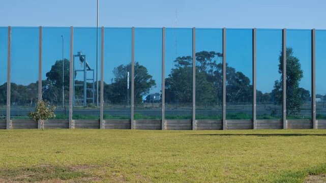 A transparent blue glass noise barrier wall alongside a highway in Melbourne, Australia, with traffic passing through the visual panel. Urban noise mitigation and visual aesthetics.