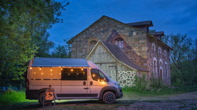 white camper van with night illumination in from of facade of old stone house in twilight