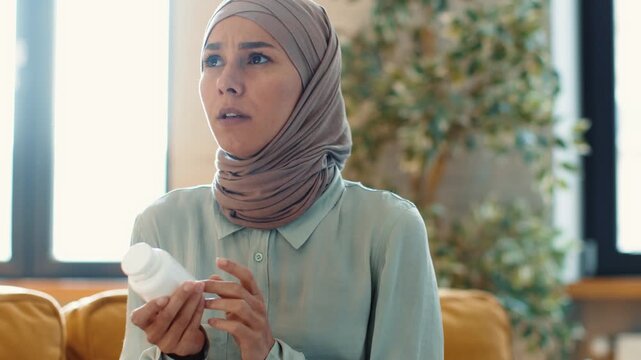 A woman with light skin, wearing a hijab, applies a skincare product while sitting in a bright living room. Plants are visible in the background adding to the home setting.
