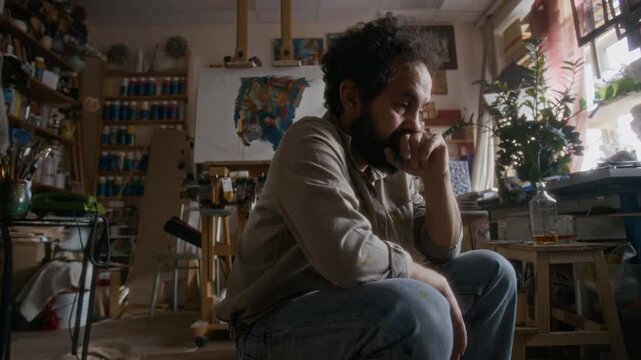 Frustrated male artist deep in thought sitting in creative studio, answering call on landline phone and having conversation