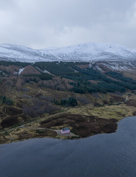 Aerial view of The Pink House on the shore of Loch Glass with snow-capped mountains and evergreen forests under a cloudy sky in Dingwall, Scotland, United Kingdom.