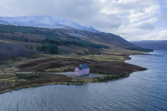 Aerial view of The Pink House on the shore of Loch Glass with snow-capped mountains and a cloudy sky in Dingwall, Scotland, United Kingdom.