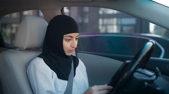 Middle Eastern woman wearing black hijab sitting in driver seat looking down at smartphone. Female driver inside modern car in daylight checking messages or navigation on mobile phone before driving.