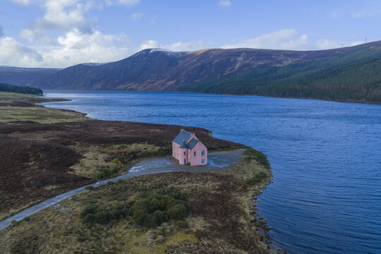 Aerial view of The Pink House on the shore of Loch Glass surrounded by rugged mountains and blue water in Dingwall, Scotland, United Kingdom.
