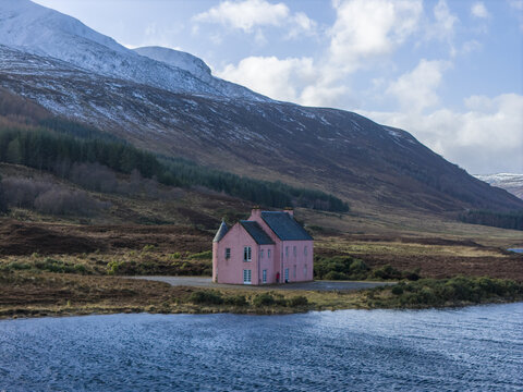 Aerial view of The Pink House on the shore of Loch Glass surrounded by snow-capped hills and pine forests Dingwall, Scotland, United Kingdom.