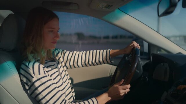 Woman in black-and-white striped sweater driving car, shifting gears with right hand while steering with left. Focused side profile, short wavy hair, sunlight casting patterns inside modern vehicle.