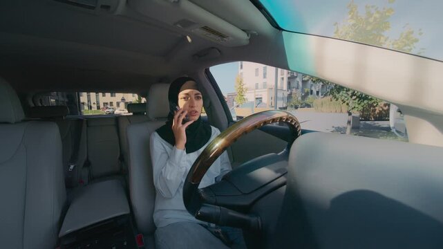 Muslim woman in black hijab sitting in driver seat holding smartphone to ear during phone call. Female talking on phone seriously inside parked car on sunny day in modern residential neighborhood.