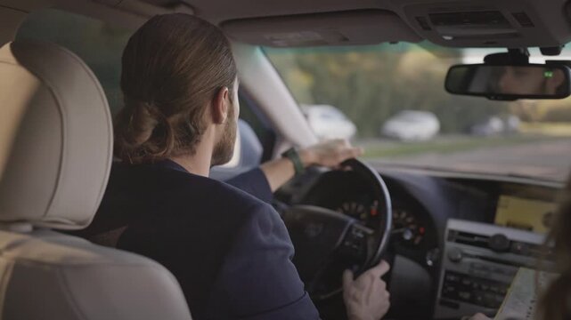 Caucasian man with beard and tied-back hair driving car, holding steering wheel and shifting gear. Wearing dark blazer, talking with passenger. Interior dashboard visible. Male driver going to work.